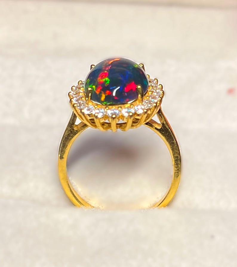 May include: A gold ring featuring a large, oval black opal gemstone with vibrant flashes of red, green, and blue. The opal is surrounded by a halo of small, clear gemstones. The ring's band is a simple, polished gold.