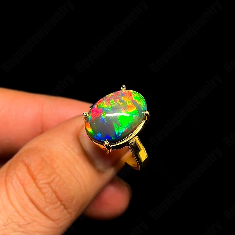 May include: A gold ring featuring a large, oval-shaped opal gemstone. The opal displays a vibrant play of color, including green, red, and blue hues. The ring's setting has four prongs securing the stone.