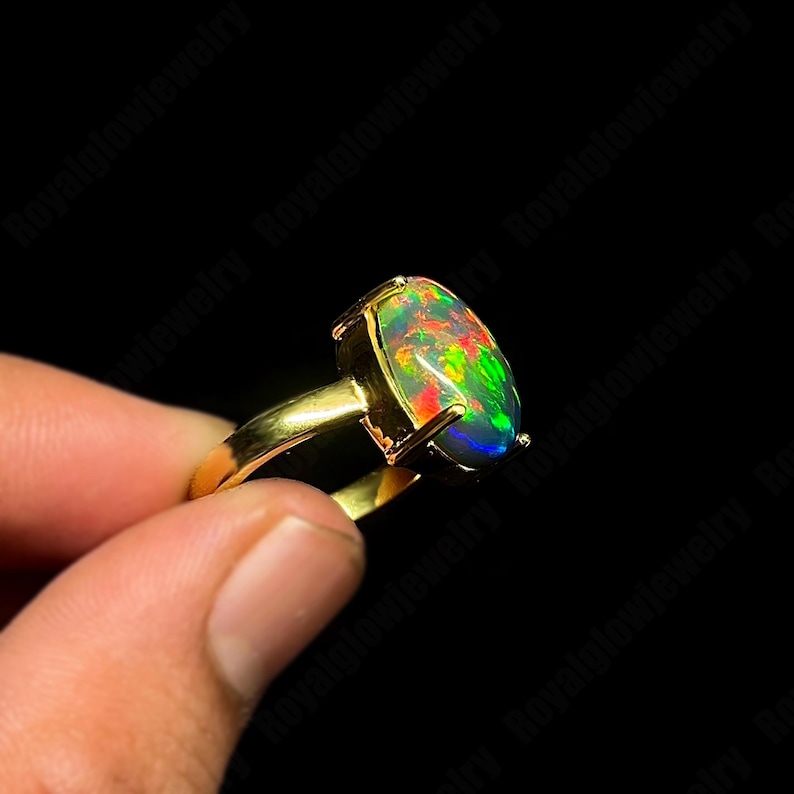 May include: A gold ring featuring a large, oval-shaped opal gemstone. The opal displays a vibrant play of color, including green, red, and blue hues. The ring's band and setting are a polished gold color, creating a striking contrast with the gemstone.