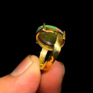 May include: A gold ring featuring an oval-shaped opal gemstone. The opal displays a mix of green, red, and black colors. The ring's band and setting are a shiny gold color, creating a striking contrast against the dark background.