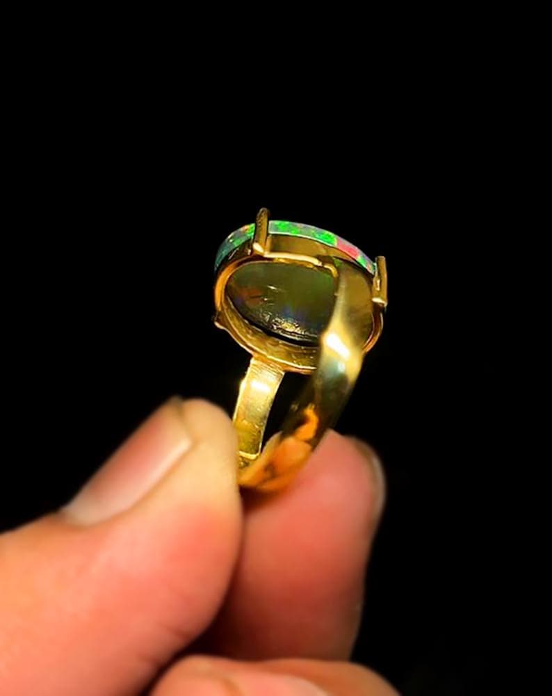 May include: A gold ring featuring an oval-shaped opal gemstone. The opal displays a mix of green, red, and black colors. The ring's band and setting are a shiny gold color, creating a striking contrast against the dark background.
