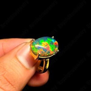 May include: A gold ring featuring a large, oval-shaped opal. The opal displays a vibrant play of color, with flashes of green, red, and yellow against a dark background. The ring's band and setting are a polished gold color.