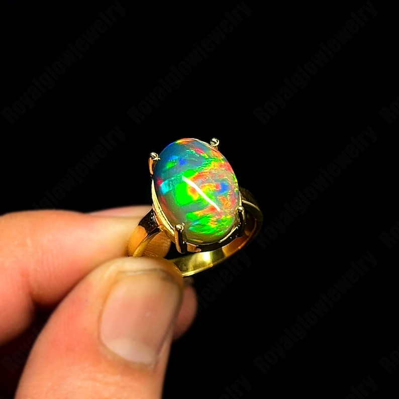 May include: A gold ring featuring an oval opal gemstone. The opal displays a vibrant play of color, including green, blue, and orange hues. The ring's band and setting are a polished gold color, creating a striking contrast with the gemstone.