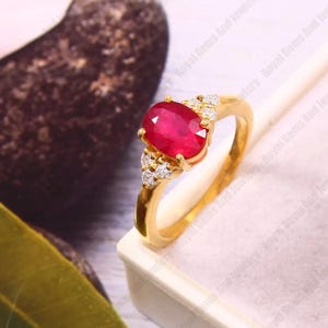 Può includere: Un anello d'oro con un grande rubino ovale rosso vivo. Piccole gemme chiare adornano il rubino. L'anello è montato su una fascia d'oro lucida.