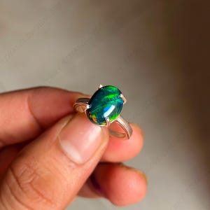 May include: A silver ring with a large, oval-cut black opal gemstone. The opal has flashes of green and blue. The ring's setting has four prongs. The ring is displayed against a neutral background. The ring is a piece of jewellery.