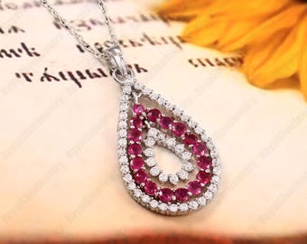 Puede incluir: Un collar con colgante de plata con un diseño de lágrima. El colgante presenta un grupo de pequeñas piedras preciosas rojas redondas rodeadas por un borde de piedras preciosas claras más pequeñas. El collar está en una delicada cadena de plata.