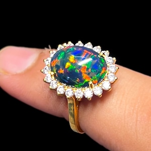 May include: A gold ring featuring a large, oval black opal gemstone with vibrant flashes of color. The opal is surrounded by a halo of small, clear gemstones. The ring's band is a simple, polished gold.