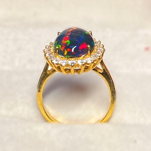 May include: A gold ring featuring a large, oval-shaped black opal gemstone with vibrant flashes of red, green, and blue. The opal is surrounded by a halo of small, clear gemstones. The ring's band is a simple, polished gold.