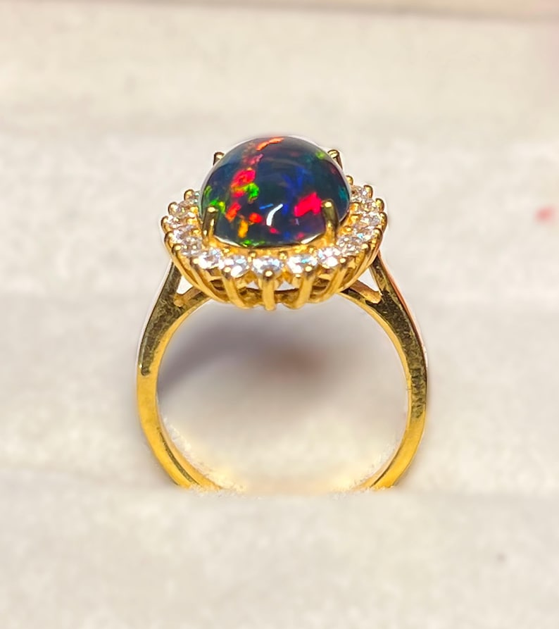 May include: A gold ring featuring a large, oval-shaped black opal gemstone with vibrant flashes of red, green, and blue. The opal is surrounded by a halo of small, clear gemstones. The ring's band is a simple, polished gold.