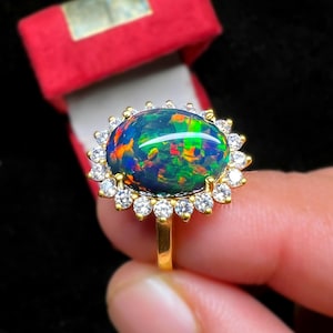 May include: A gold ring featuring a large, oval black opal gemstone with vibrant internal colors. The opal is surrounded by a halo of small, clear gemstones. The ring is held in front of a red jewelry box.