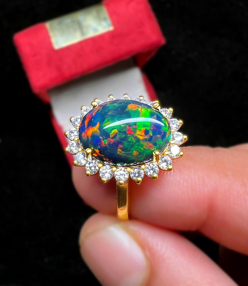 May include: A gold ring featuring a large, oval black opal gemstone with vibrant internal colors. The opal is surrounded by a halo of small, clear gemstones. The ring is held in front of a red jewelry box.
