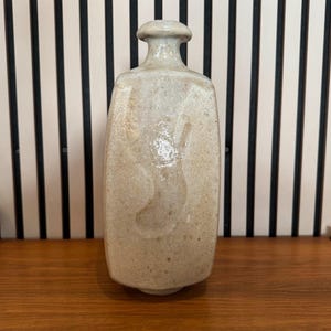 Könnte beinhalten: Eine beige Keramikflasche mit rundem Deckel und rechteckigem Körper. Die Flasche hat eine strukturierte Oberfläche und einen kleinen, kreisförmigen Verschluss. Die Flasche steht auf einer Holzoberfläche.