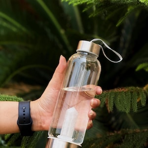 Puede incluir: Una botella de agua de vidrio transparente con tapa y base plateadas, llena de agua y un cristal blanco. La botella se sostiene en una mano, con un reloj negro visible en la muñeca. El fondo es follaje verde borroso.