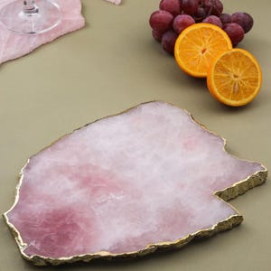 May include: A pink, irregularly shaped serving tray with a gold-coloured edge. The tray is made of a natural stone material and is displayed with sliced oranges and grapes. The tray is ideal for serving appetizers or displaying decorative items.