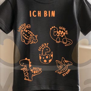 Puede incluir: Una camiseta negra con gráficos de dinosaurios naranjas y la frase "ICH BIN" en la parte superior. Debajo, varias ilustraciones de dinosaurios con palabras en alemán. La camiseta está colgada en un perchero de madera.