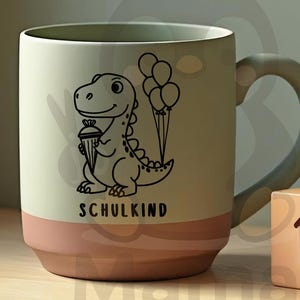 May include: A ceramic mug with a light green upper half and a pink lower half. A black cartoon dinosaur holding a cupcake and balloons is printed on the mug. The word "SCHULKIND" is printed below the dinosaur. A small cube with the number 15 is next to the mug.