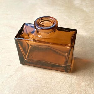 May include: A square amber glass bottle with a cylindrical neck. The bottle has a geometric design with angled sides and a flat base. The glass is transparent, allowing light to pass through. The bottle is empty.