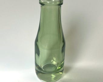 May include: A clear, pale green glass bottle with a rounded base and a narrow neck. The bottle is empty and has a smooth surface. The bottle is likely used for storing liquids.