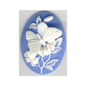May include: A white ceramic cameo with a butterfly design on a light blue background. The butterfly is in flight and has detailed wings. The cameo is oval shaped.