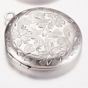 以下が含まれることがあります： 花柄と渦巻き模様の銀色の丸いロケット。ロケットはヒンジ付きの開口部があり、チェーンに取り付けるための小さなループが上部にあります。 デザインは表面にエンボス加工されています。