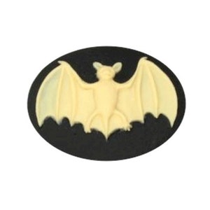 May include: A cream-colored bat with outstretched wings is centered on a black oval. The bat is raised, creating a 3D effect. The image is a decorative element, possibly for Halloween or gothic-themed crafts.