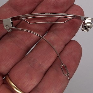 May include: Close-up of two silver-colored metal hair barrettes. One is open, showing the spring mechanism and the curved metal piece that holds the hair. The other is closed. The barrettes are held in a hand.