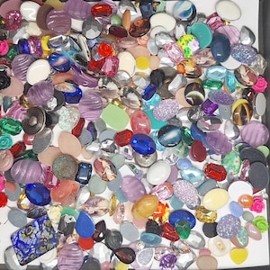 May include: A collection of assorted cabochons and flat-back embellishments in various shapes, sizes, and colors, including translucent, opaque, glittered, and floral-patterned pieces, scattered together in a square container.