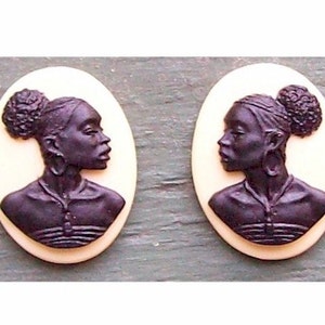 May include: Two black cameo pendants with a cream background. Each pendant features a profile of a woman with a bun in her hair. The woman is wearing a necklace and a top.