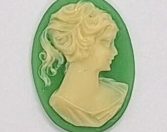 May include: Oval cameo pendant featuring a profile of a woman with flowing hair and a draped garment. The woman's image is cream-colored, set against a green background. The cameo is likely made of resin or a similar material.