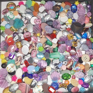 May include: A collection of assorted craft embellishments including colorful cabochons, faceted acrylic gems, small plastic flowers, buttons, and oval cameos with printed portraits, all mixed together in a square container.