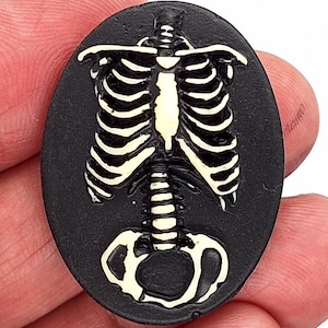 May include: An oval-shaped, black pendant featuring a detailed, off-white skeleton torso design. The rib cage, spine, and pelvis are clearly defined against the dark background. This gothic-themed pendant is suitable for jewelry making or crafting.