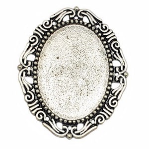 May include: An oval-shaped, silver-toned metal jewelry finding with an ornate, filigree-style border. The center is a textured, oval surface, ready for embellishment. The decorative frame features scrollwork and beaded accents, perfect for crafting.