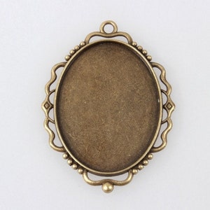 May include: An antique bronze-colored oval pendant tray with a decorative frame. The frame features a scalloped edge, small beaded accents, and a loop at the top for hanging. The pendant tray is designed for jewelry making.