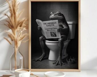 May include: A framed black and white wall art print featuring a toad sitting on a toilet reading a newspaper titled The Amphibian Gazette with the headline Local Toad Claims He's a Prince, with a small crown resting on the floor beside the toilet.