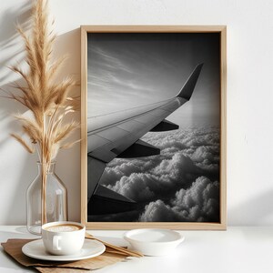 May include: A black and white framed wall art print depicting an airplane wing soaring above a dense layer of clouds. The rectangular wooden frame holds the monochromatic photograph, which captures the structural details of the wing in flight.