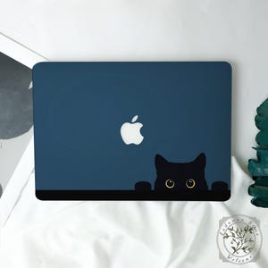 Könnte beinhalten: Ein marineblauer Laptop mit weißem Apple-Logo und einem schwarzen Katzenmotiv. Der Kopf und die Pfoten der Katze schauen über einen schwarzen Rand, mit gelben Augen. Der Laptop liegt auf einer weißen Oberfläche.