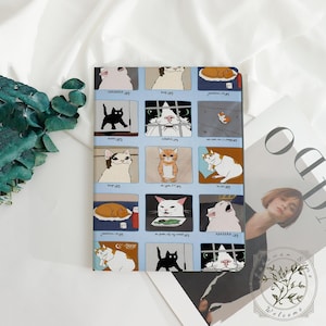 May include: A rectangular notebook featuring a light blue background with a grid of cat illustrations. Each cat has a different expression. The notebook is lying on a white surface with a green plant and a magazine in the background.