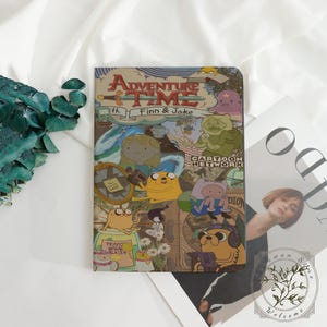 May include: A notebook featuring a collage of Adventure Time characters and the show's logo. The cover has a tan background with images of Finn, Jake, and other characters from the Cartoon Network show. The words "Adventure Time with Finn & Jake" are displayed at the top.
