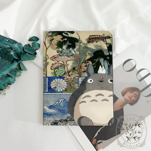 Peut inclure: Un carnet avec une illustration colorée de Totoro, un esprit de la forêt gris, avec une scène forestière fantaisiste et une bordure à motifs bleus et blancs. Le carnet est rectangulaire avec des coins arrondis.