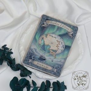 May include: A vintage-style journal with a cover featuring a world map, a sailing ship, and an aurora borealis design. The words "Sophia Edition" and "Journal" are displayed on the cover. The journal is resting on a white, decorative plate.