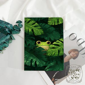 Op de afbeelding: Een rechthoekig notitieboek met een levendige illustratie van een groene kikker met grote ogen die achter dichte, donkergroene tropische monstera-bladeren vandaan kijkt. De kaft toont een weelderig oerwoudthema met gelaagd gebladerte.