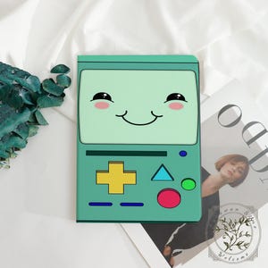 May include: A teal-colored case featuring a cartoon character with a smiling face, rosy cheeks, and black eyes. The case has a yellow cross, a blue triangle, and a red circle. The case is on a white surface.
