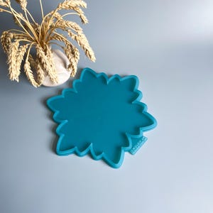 Pu&ograve; includere: Uno stampo in silicone turchese a forma di fiore. Lo stampo ha un bordo smerlato e si trova su uno sfondo azzurro. Sullo sfondo c'&egrave; un piccolo vaso bianco con spighe di grano.