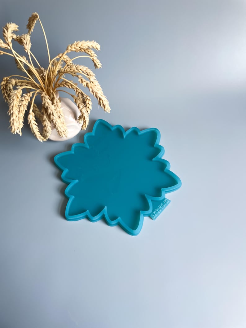 Pu&ograve; includere: Uno stampo in silicone turchese a forma di fiore. Lo stampo ha un bordo smerlato e si trova su uno sfondo azzurro. Sullo sfondo c'&egrave; un piccolo vaso bianco con spighe di grano.