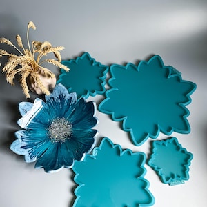 Pode incluir: Uma coleção de moldes de resina turquesa em forma de flor em vários tamanhos, juntamente com uma tigela de flor de resina acabada com um design marmorizado azul e branco e um centro de glitter prateado. Um pequeno vaso com trigo seco também está presente.