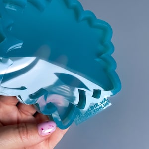 Pu&ograve; includere: Uno stampo in silicone traslucido color turchese a forma floreale. Lo stampo &egrave; tenuto in una mano con una manicure rosa chiaro. Lo stampo ha una superficie liscia e lucida ed &egrave; progettato per l'artigianato.