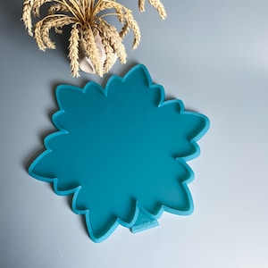 Pu&ograve; includere: Uno stampo in silicone turchese a forma di fiore. Lo stampo ha un bordo smerlato e una superficie liscia. L'immagine mostra anche un piccolo vaso con spighe di grano.