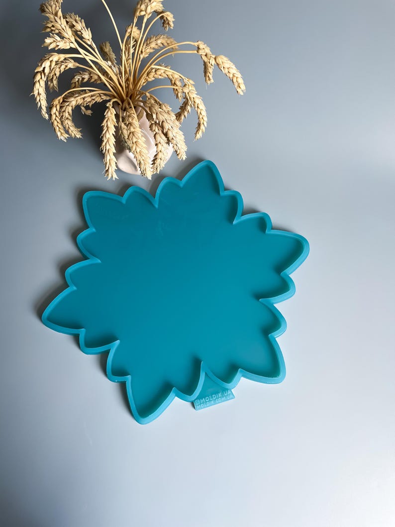 Pu&ograve; includere: Uno stampo in silicone turchese a forma di fiore. Lo stampo ha un bordo smerlato e una superficie liscia. L'immagine mostra anche un piccolo vaso con spighe di grano.