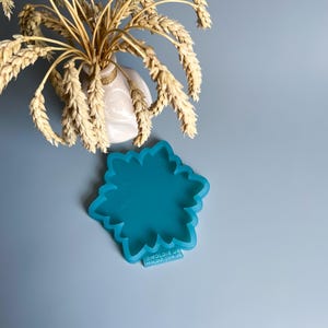 Pu&ograve; includere: Uno stampo in silicone turchese a forma di fiore, con un bordo smerlato, &egrave; posto su una superficie azzurra. Un piccolo vaso bianco con spighe di grano &egrave; sullo sfondo. Lo stampo ha il testo "@MOLDIK.UA MOLDIK.COM.UA".