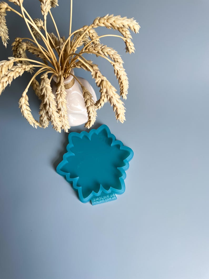 Pu&ograve; includere: Uno stampo in silicone turchese a forma di fiore, con un bordo smerlato, &egrave; posto su una superficie azzurra. Un piccolo vaso bianco con spighe di grano &egrave; sullo sfondo. Lo stampo ha il testo "@MOLDIK.UA MOLDIK.COM.UA".